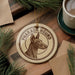 A circular ornament featuring a hound dog's head, with the text "PHARAO HOUND" printed on it, is displayed on a wooden surface alongside a cup of coffee and some pine branches.