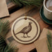 a round ornament with a bird illustration on it, placed on a wooden surface next to a cup of coffee and some pine branches.
