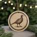 A round ornament featuring a bird, possibly a pheasant, is displayed in front of a blurred Christmas tree with twinkling lights.