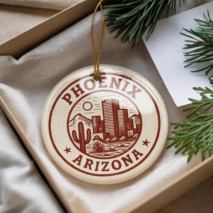 A decorative ornament with the text "PHOENIX ARIZONA" printed on it, hanging from a string. The ornament is placed on a white surface, with some greenery visible in the background.