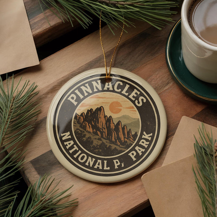 A circular ornament with the text "PINNACLES NATIONAL PARK" and an illustration of mountains is displayed on a wooden surface, surrounded by pine branches and a cup of coffee.