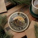 A circular ornament with the text "PINNACLES NATIONAL PARK" and an illustration of mountains is displayed on a wooden surface, surrounded by pine branches and a cup of coffee.