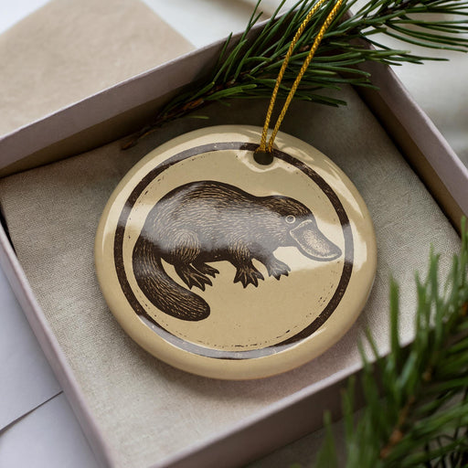 A ceramic ornament in the shape of a platypus, with a brown and beige color scheme, is displayed in a box with pine branches.