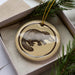 A ceramic ornament in the shape of a platypus, with a brown and beige color scheme, is displayed in a box with pine branches.