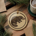 a round ornament with a black and white illustration of a platypus on a wooden surface, surrounded by pine branches and a cup of coffee.