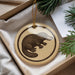 A round ornament featuring a black and brown illustration of a platypus is displayed in a box, surrounded by pine branches.