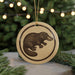 A round ornament featuring a black illustration of a platypus is displayed in front of a blurred background of a Christmas tree with lights.