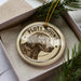 A circular ornament with a dog's head and the text "PLOTT HOUND" printed on it, placed inside a box with pine branches.