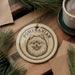 A ceramic ornament featuring a Pomeranian dog's face, with the word "POMERANIAN" written around the image. The ornament is placed on a wooden surface, with pine branches and a cup of coffee nearby.