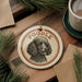 A circular ornament featuring a poodle's portrait is displayed on a wooden surface, accompanied by a cup of coffee and pine branches.