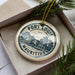 A decorative ornament in a box, featuring a circular design with text and an illustration of a building and mountains.