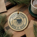 a round ornament with the text "PORTUGUESE MAN O' WAR" on it, surrounded by pine branches and a cup of coffee.