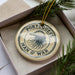 A circular ornament with the text "PORTUGUESE MAN O' WAR" printed on it, surrounded by a pine branch and pine needles.