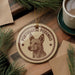 A round ornament featuring a dog's head and the text "PORTUGUESE PODENGO" is displayed on a wooden surface, surrounded by pine branches and a cup of coffee.