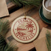 A circular ornament with the text "PRASLIN SEYCHELLES" is displayed on a wooden surface, surrounded by pine branches and a cup of coffee.