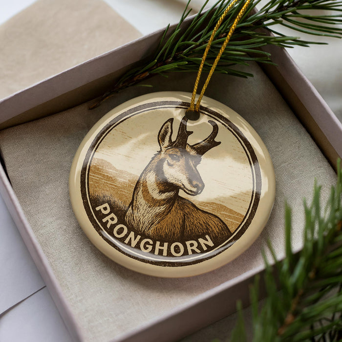 A circular ornament with a deer image and the word "PRONGHORN" printed on it, placed in a box with pine branches.