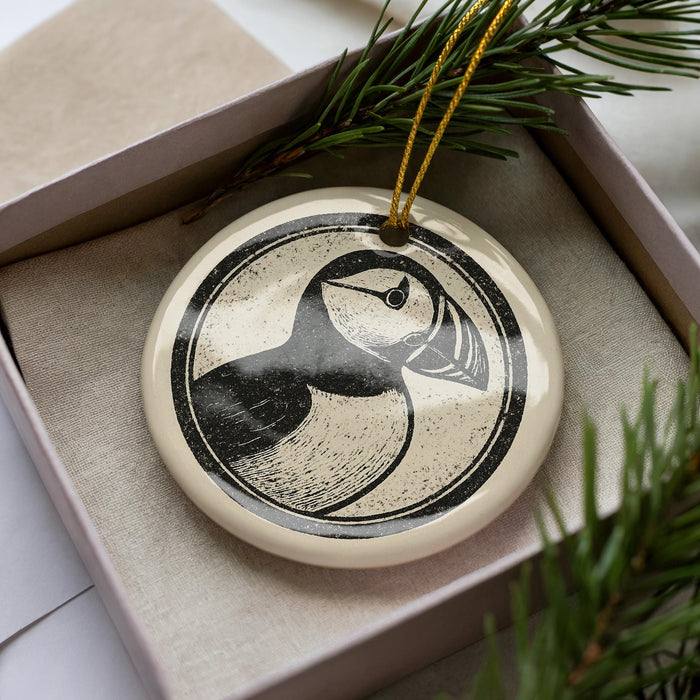 A decorative ornament in a box, featuring a black and white illustration of a bird, likely a puffin, with a yellow string hanging from it.
