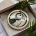 A decorative ornament in a box, featuring a black and white illustration of a bird, likely a puffin, with a yellow string hanging from it.