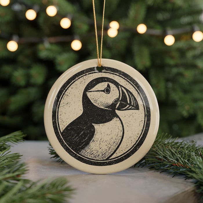 A circular ornament featuring a black and white illustration of a puffin bird is displayed against a blurred background of Christmas lights.