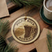 A circular ornament with the text "PULPIT ROCK NORWAY" on it, featuring a scenic image of a cliff with people standing on it. The ornament is hanging from a string, and there is a cup of coffee and some pine branches in the background.