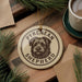 A circular ornament featuring a dog's face and the text "PYRENEAN SHEPHERD" is displayed on a wooden surface, surrounded by pine branches and a cup of coffee.