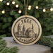 A circular ornament with a drawing of a church and the text "PEÇS HUNGARY" on it, hanging in front of a Christmas tree with lights.