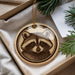 A round ornament featuring a raccoon's face is displayed in a box, surrounded by pine branches.