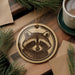 a round ornament with a raccoon's face on it, placed on a wooden surface next to a cup of coffee and some pine branches.
