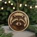 A round ornament featuring a raccoon's face is displayed in front of a blurred background of Christmas lights.