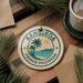 A circular ornament with the text "RANGIRO A FENCH POLYNESIA" and a tropical beach scene, hanging on a wooden surface with pine branches and a cup of coffee nearby.