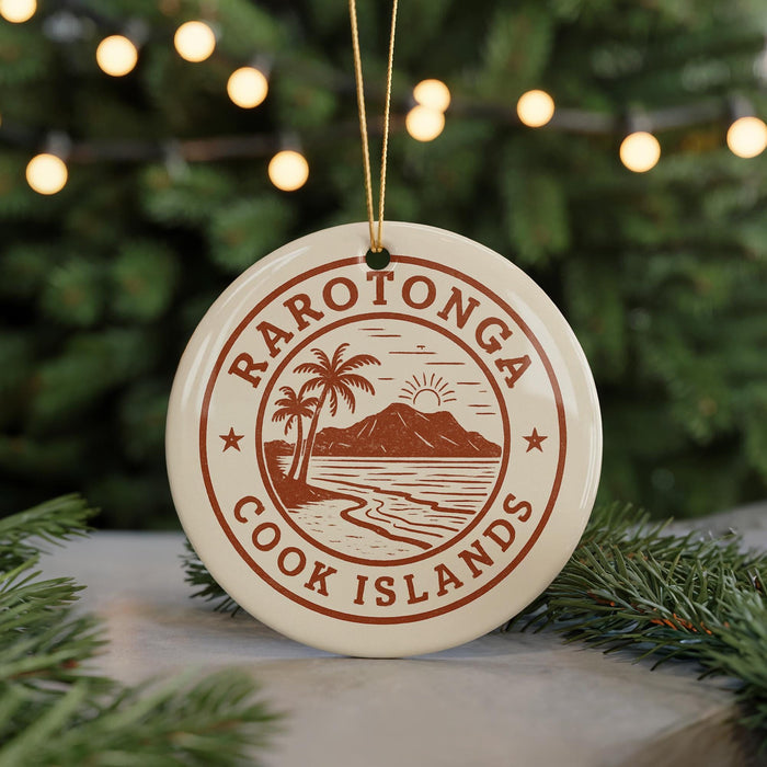 A circular ornament with the text "RAROTONGA COOK ISLANDS" and a graphic of a beach and palm trees, hanging from a string against a background of Christmas lights and greenery.