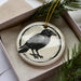 A ceramic ornament in the shape of a black bird is displayed in a box, surrounded by pine branches.