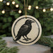 A ceramic ornament featuring a black crow silhouette is displayed against a blurred background of Christmas lights.