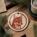 a red and white ceramic ornament with a fox's face on it, hanging on a wooden surface. There is also a cup of coffee and some pine branches in the background.