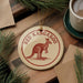 A round ornament with a kangaroo image and the text "RED KANGAROO" is displayed on a wooden surface, surrounded by pine branches and a cup of coffee.