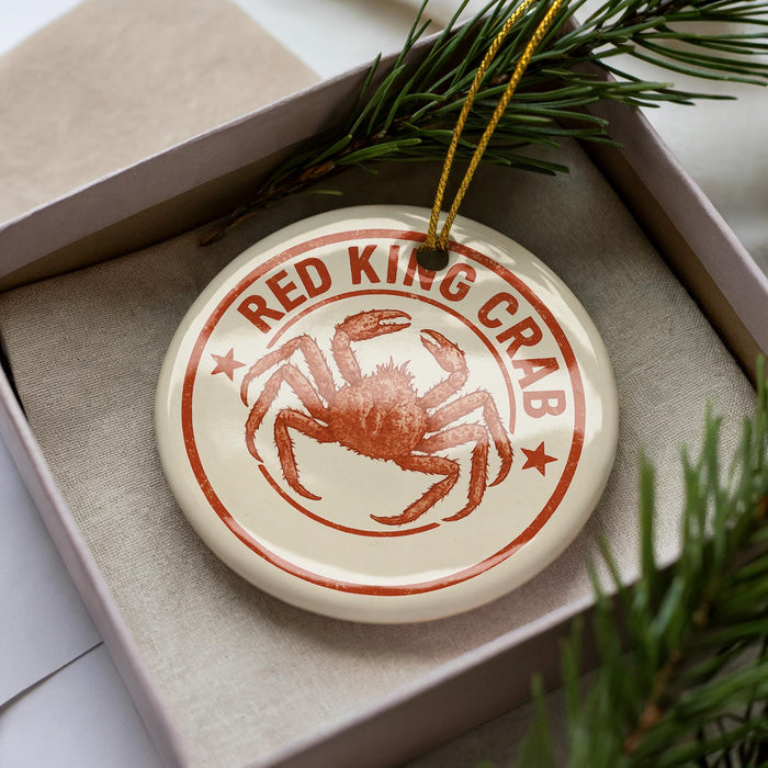 A round ornament with a red crab design, placed inside a box with pine branches.