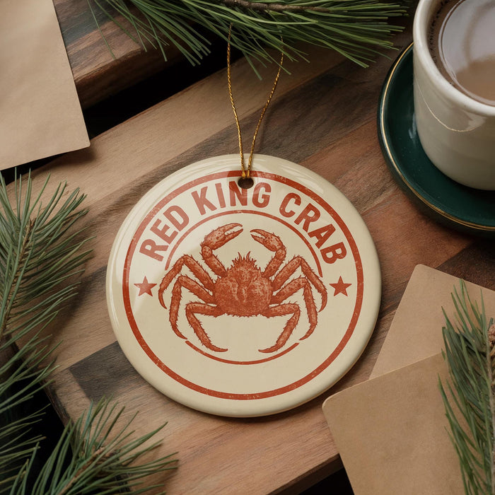 A round ornament featuring a red crab is displayed on a wooden surface, surrounded by pine branches and a cup of coffee.