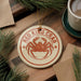 A round ornament featuring a red crab is displayed on a wooden surface, surrounded by pine branches and a cup of coffee.