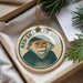 A round ornament featuring a monkey with a humorous caption, "RED-SHANKED DOUC", is displayed in a box with pine branches.
