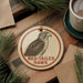 A round ornament featuring a red-tailed hawk is displayed on a wooden surface, surrounded by pine branches and a cup of coffee.