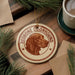 A circular ornament featuring a brown dog's head, with the text "REDBONE COONHOUND" around the edge, placed on a wooden surface next to a cup of coffee and pine branches.