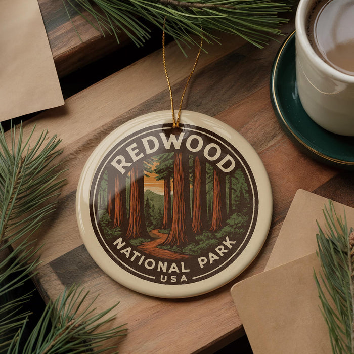 A circular ornament with the text "REDWOOD NATIONAL PARK USA" and an illustration of a forest of tall trees, placed on a wooden surface next to a cup of coffee and some pine branches.
