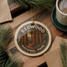 A circular ornament with the text "REDWOOD NATIONAL PARK USA" and an illustration of a forest of tall trees, placed on a wooden surface next to a cup of coffee and some pine branches.