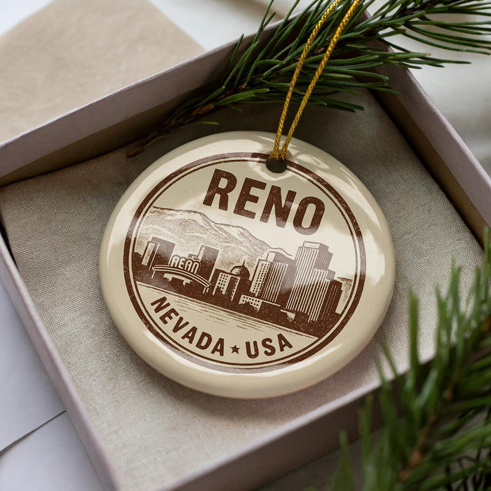 A decorative ornament with the word "Reno" and " Nevada + USA" printed on it, placed inside a box with pine branches.