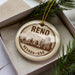 A decorative ornament with the word "Reno" and " Nevada + USA" printed on it, placed inside a box with pine branches.