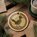 a ceramic ornament with a bird illustration, placed on a wooden surface alongside a cup of coffee and some pine branches.