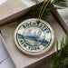 A decorative ornament in a box, featuring a circular design with a cityscape and the text "Rochester, New York" printed on it.