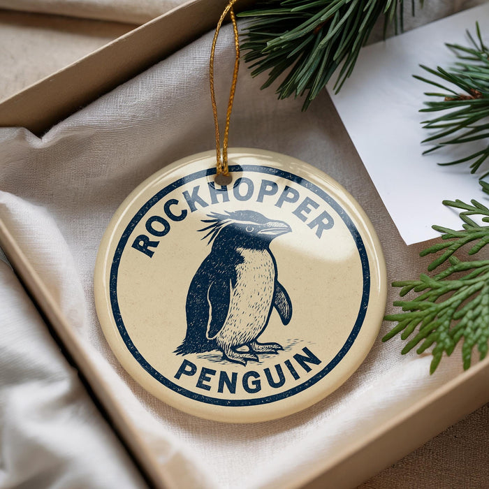 A round ornament with a penguin design, hanging from a string, placed inside a box with pine branches.