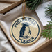 A round ornament with a penguin design, hanging from a string, placed inside a box with pine branches.