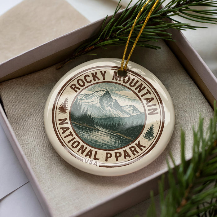 A decorative ornament in a box, featuring a mountain landscape and pine trees, with the text "ROCKY MOUNTAIN NATIONAL PARK USA" printed on it.